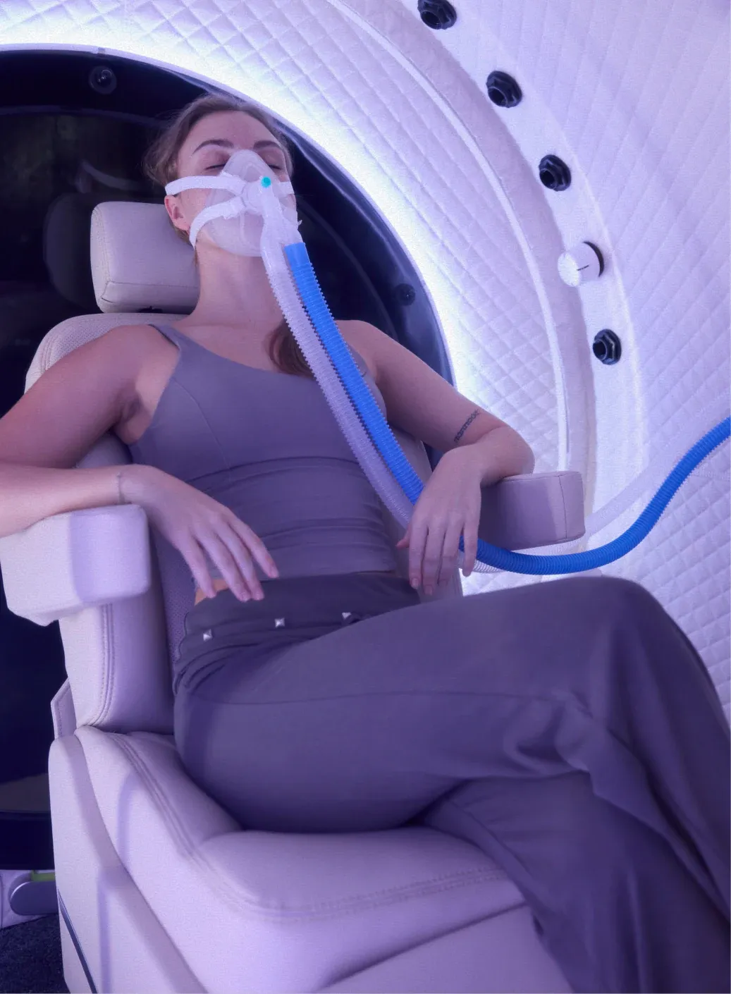 A woman reclined in the seat of a Superhuman chamber, wearing an oxygen mask