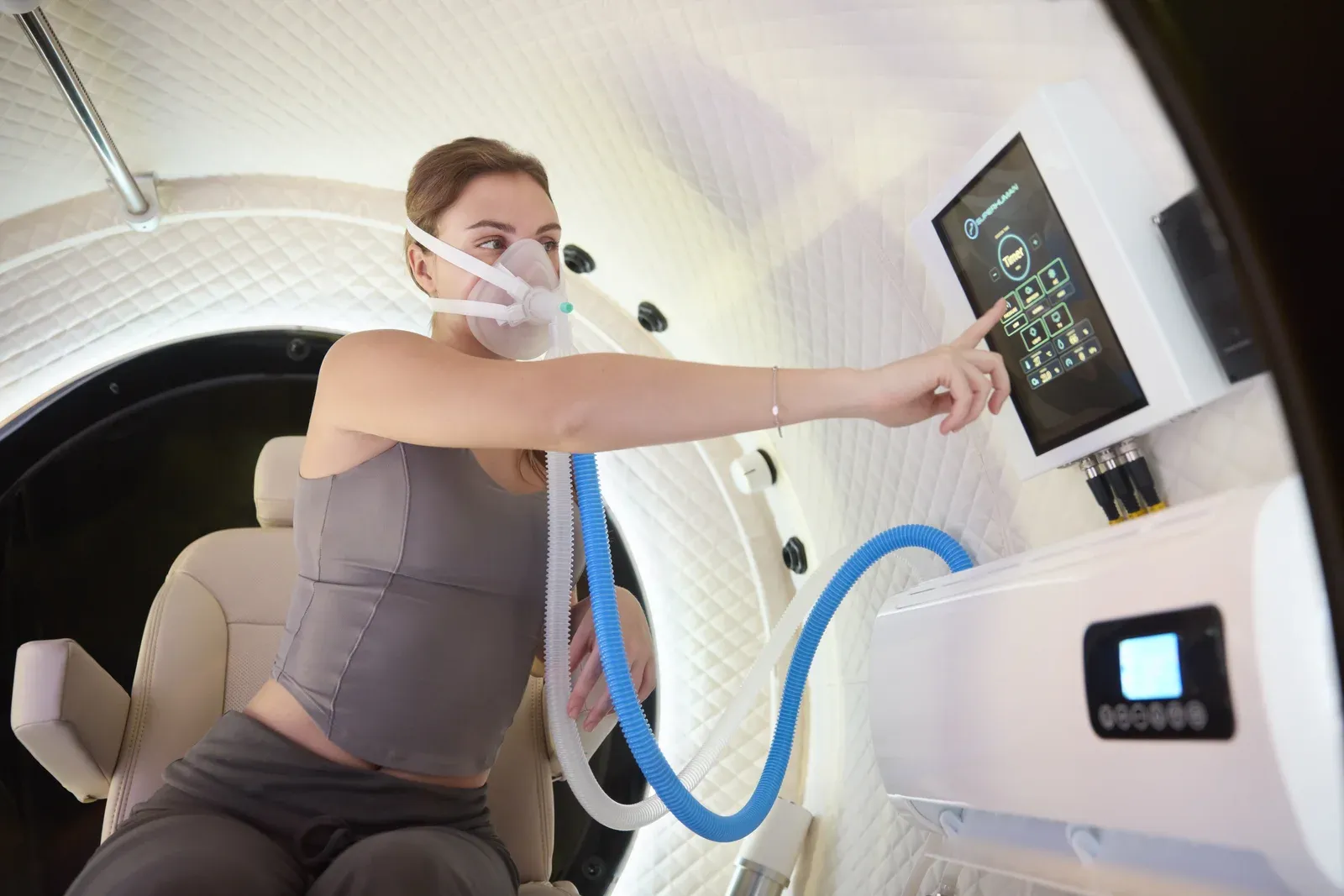 Woman breathing oxygen through a mask inside a Superhuman hard-shell hyperbaric chamber, adjusting the touchscreen