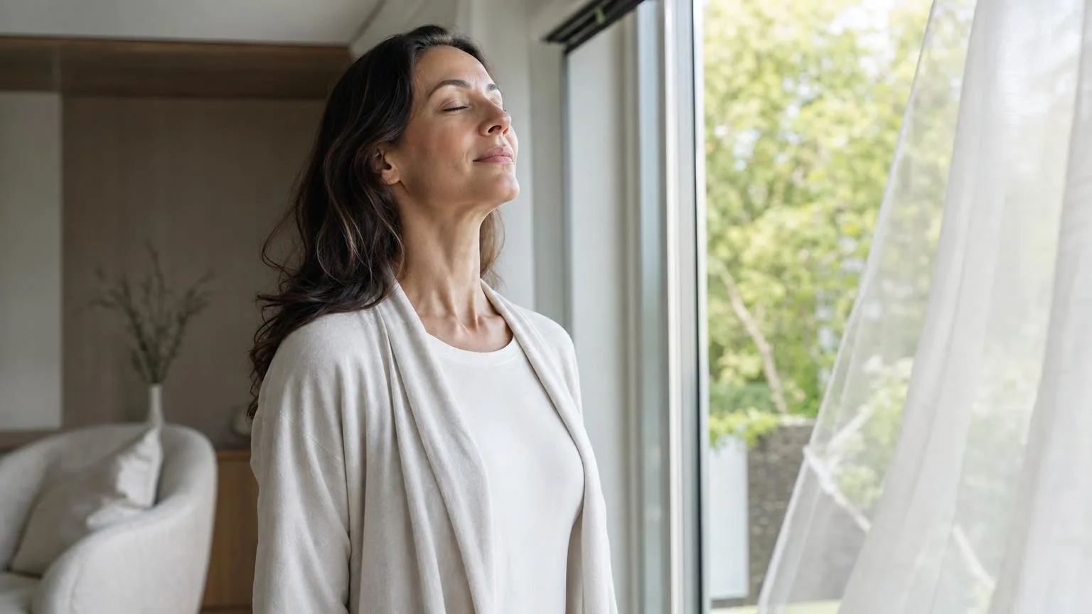 Woman taking a slow deep breath by an open window with sheer curtains and green trees outside