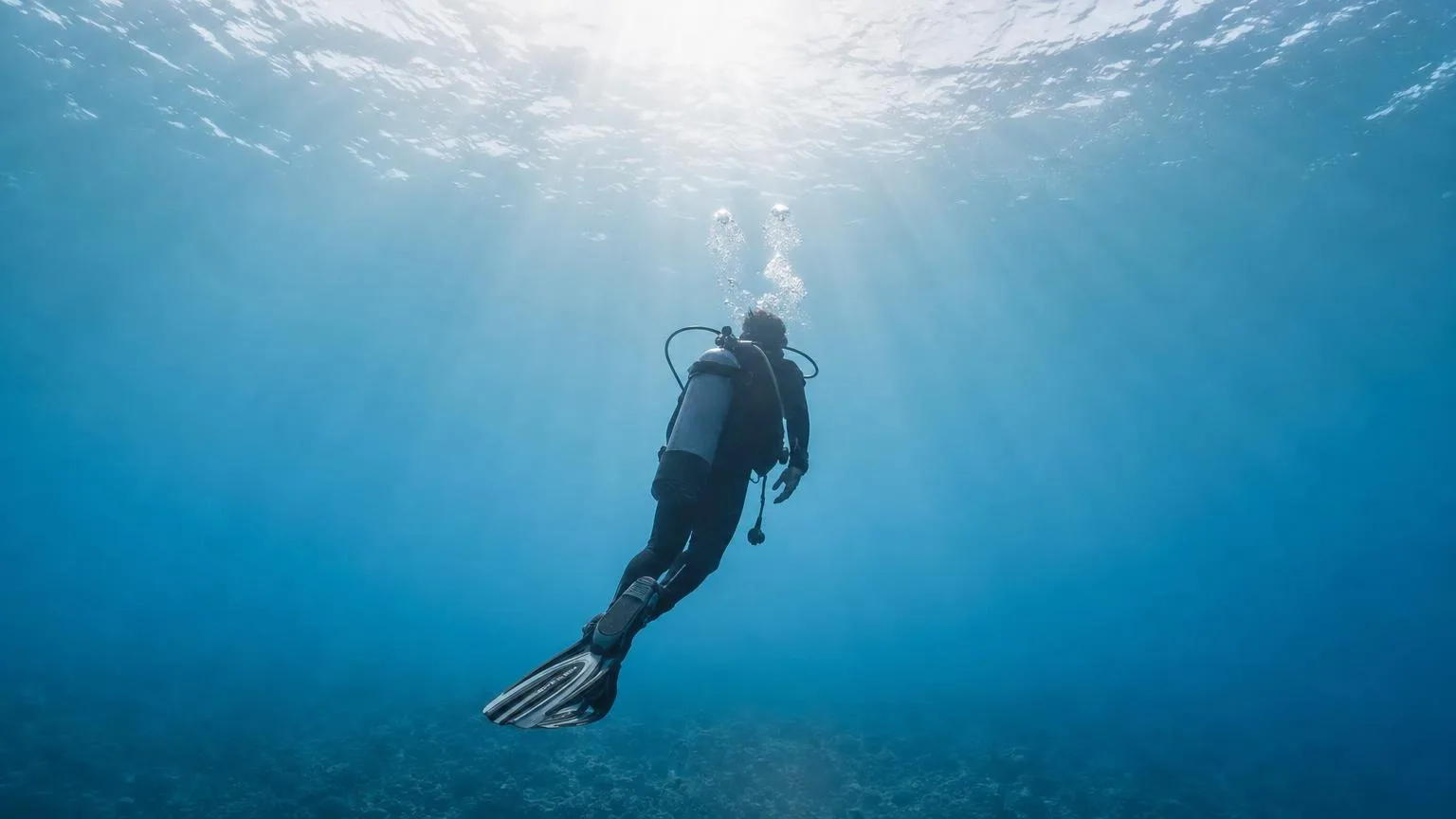 Lone scuba diver ascending slowly through calm blue water toward a bright surface
